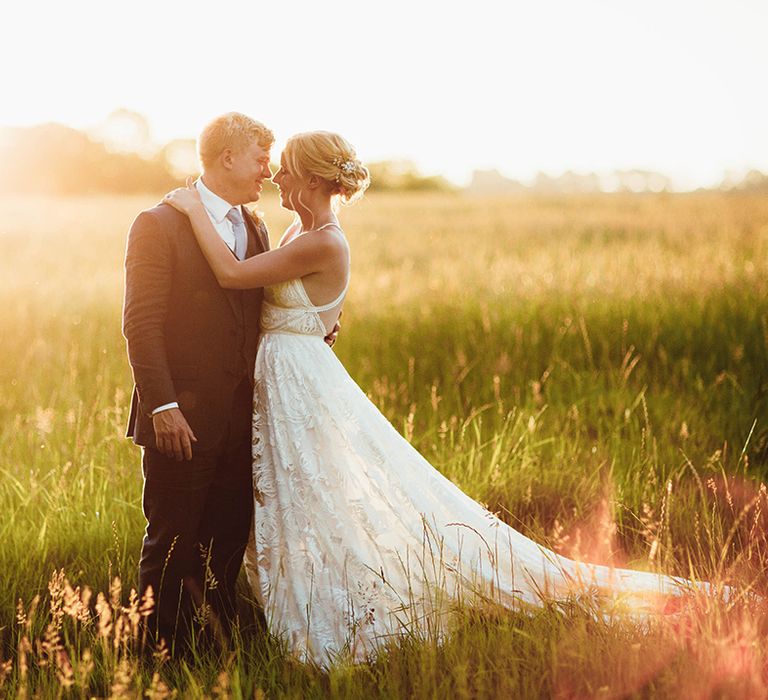 Bride and groom hug at golden hour in a field together with open back style lace wedding dress