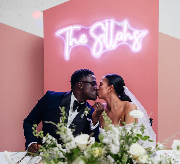 Bride and groom in classic wedding outfits sign their wedding register in front of white neon sign with the couple's new last name