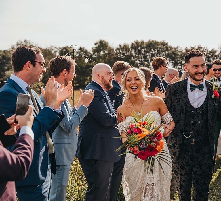 Bride and groom walk through rows of wedding guests celebrating their marriage followed by groomsmen in green suits and black bow ties