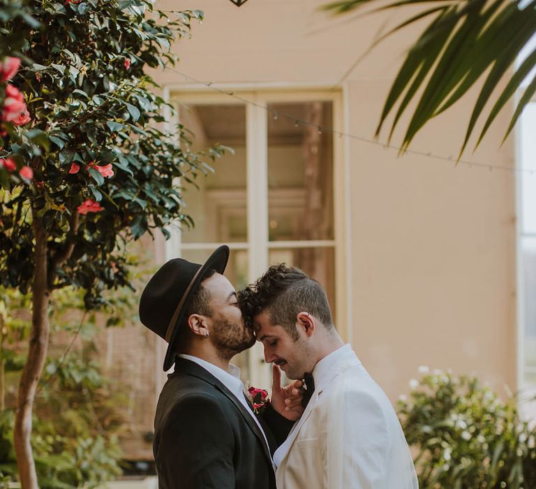 Groom in a black suit and fedora hat kissing his partner head in a white groom suit and cape