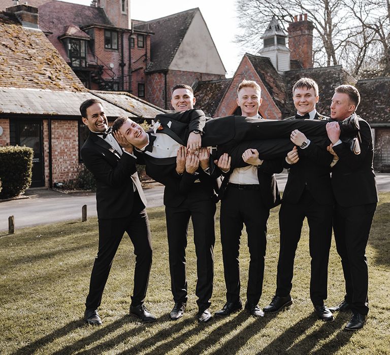 Groomsmen all hold the groom sideways wearing black tie outfits for Burley Manor wedding 