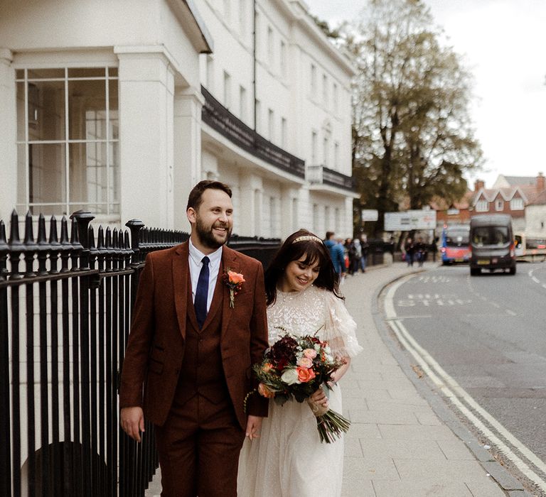 Groom in brown suit and blue tie smiles as he walks with the bride in a golden headband for intimate wedding 