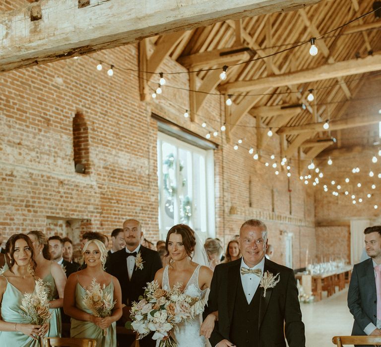 Father of the bride wearing gold bow tie and dried flower buttonhole walks the bride down the aisle at Godwick Barn