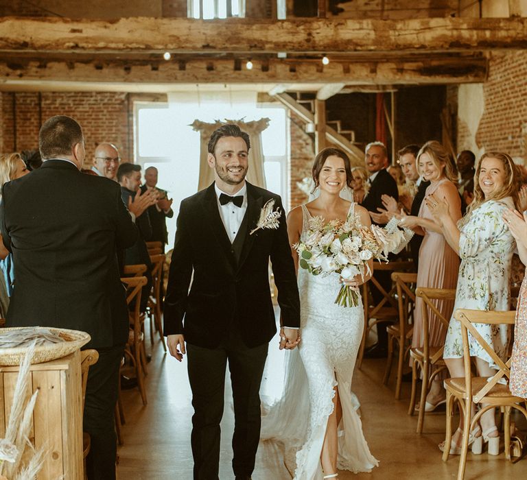 Bride and groom exit their barn wedding ceremony with groom in black tie and bride in lace wedding dress