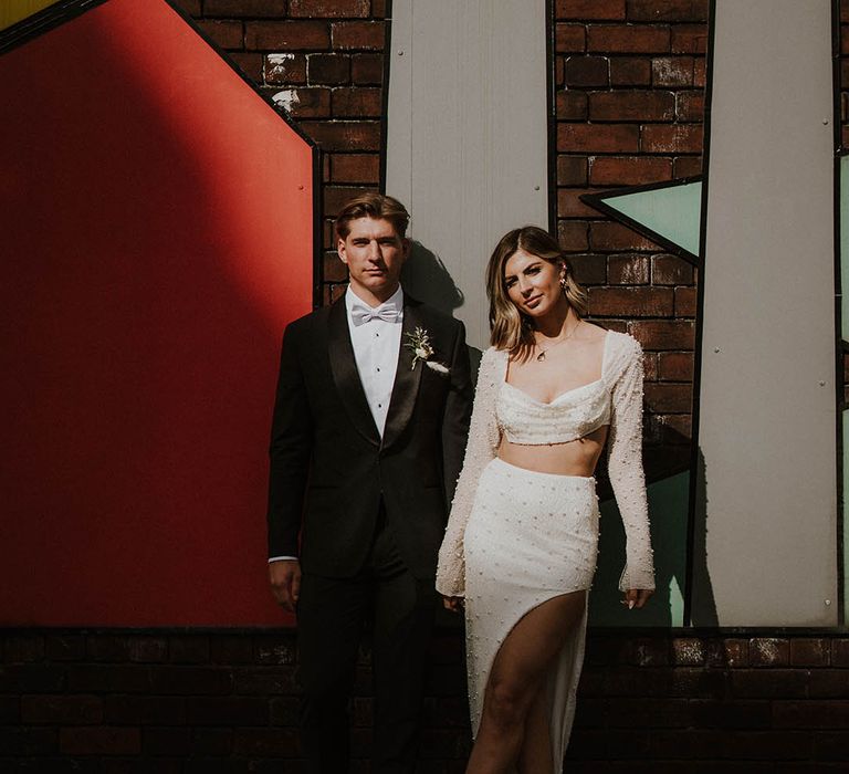 Bride and groom pose together with groom in suit jacket with satin lapels and pale blue bow tie and bride in two piece pearl outfit and white feather shoes