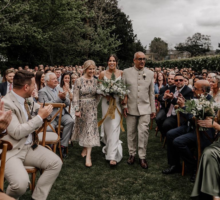 Mother and father of the bride walking their daughter down the aisle at her outdoor back garden wedding 