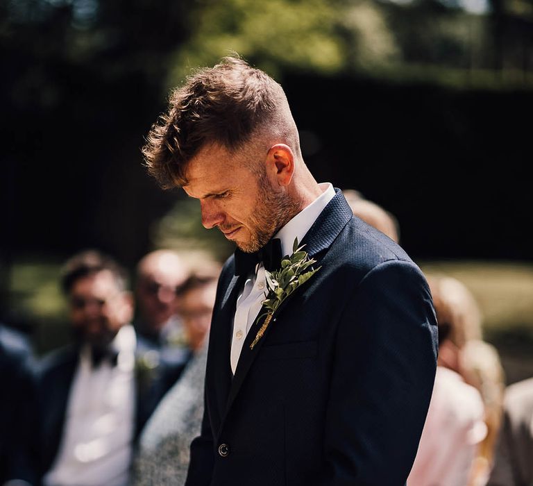 Groom waits patiently for the bride in blue tuxedo with black bow tie 