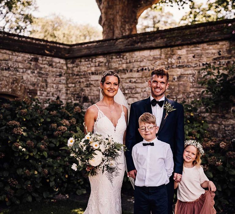 Bride and groom stand holding hands with their son and daughter for outdoor wedding ceremony 