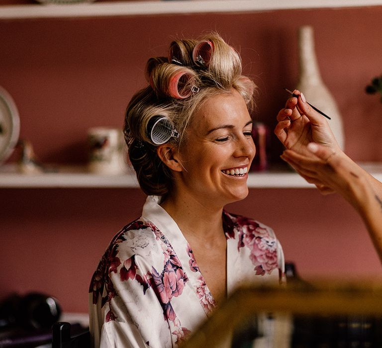 Bride in pink and white floral satin robe gets her makeup done for the wedding day with her hair in rollers 