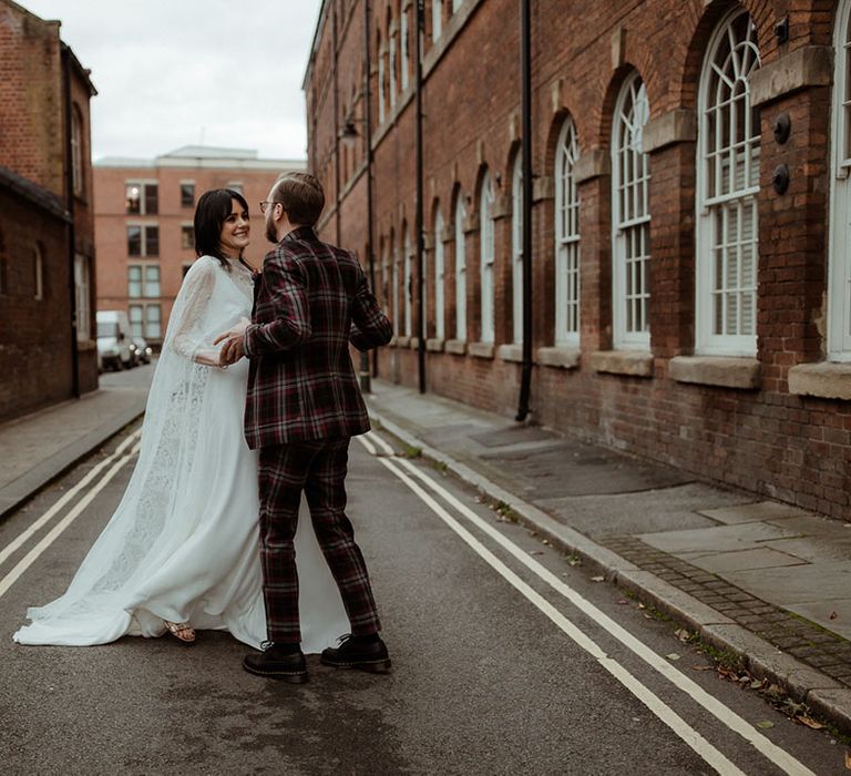 Bride & groom walk through industrial street as bride wears lace cape and groom wears bespoke tartan suit 