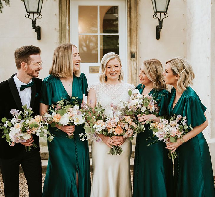 Bride stands with her bridesmaids who wear emerald green satin bridesmaid dresses with short sleeves and Man of Honour in black tie with matching green bow tie 