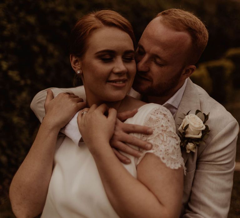 Groom holds his arm around bride who wears simple gown with lace detailing to sleeves 