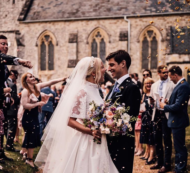 Bride in floral lace wedding dress and floral headband smiles with the groom as they have their confetti moment 