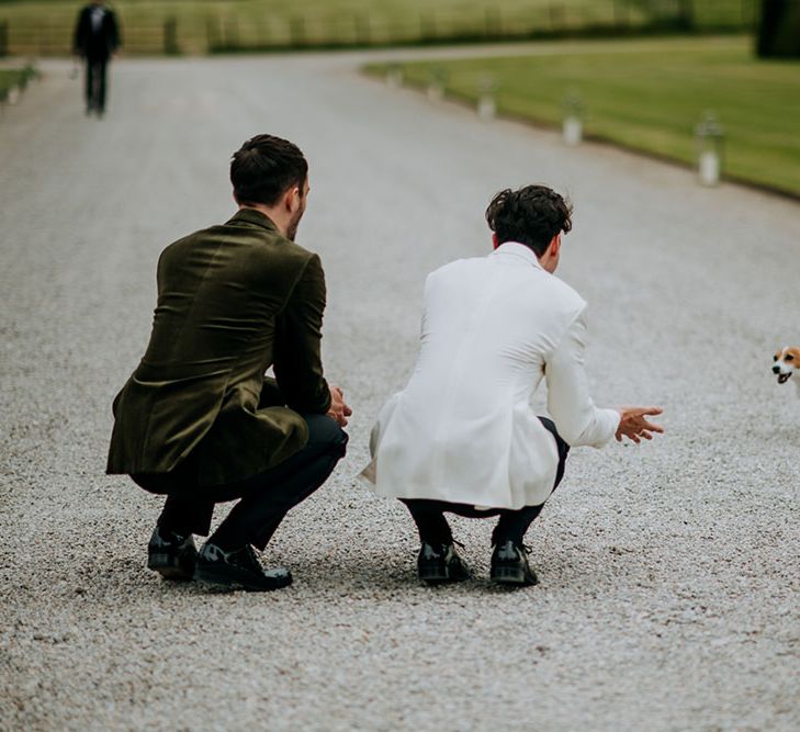 Grooms kneel on the ground to greet their Jack Russell outdoors on their wedding day