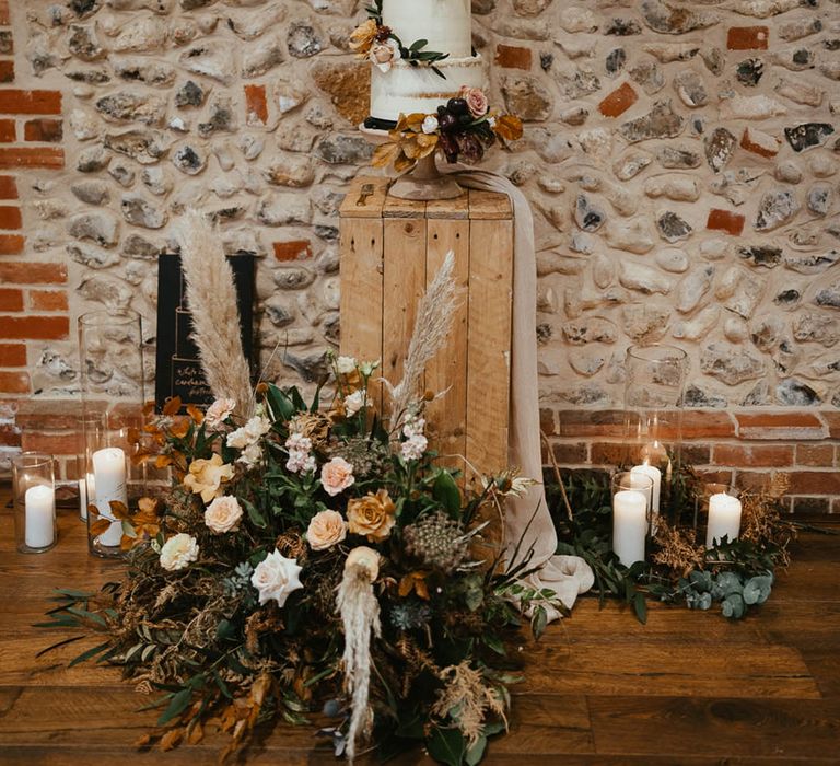 Four-tier white frosted rustic wedding cake with autumnal flowers and a personalised cake topper on a wooden crate with candles 