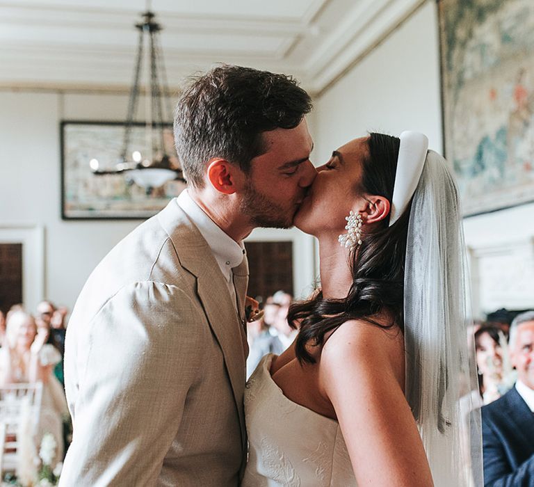 Bride wearing statement pearl earrings and groom in neutral suit share their first kiss as a married couple 