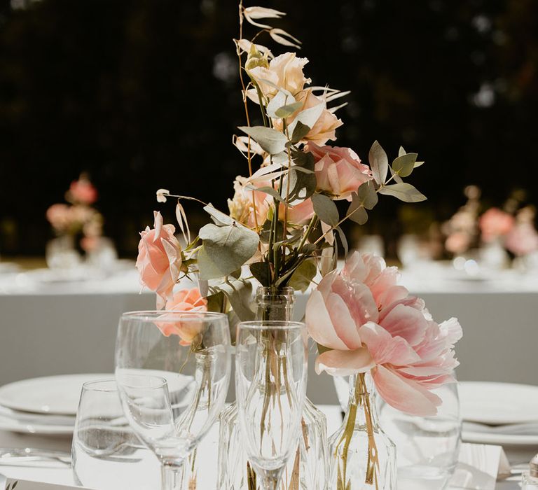 Pale pink peonies and coral florals in small glass vases across tables for outdoor reception 