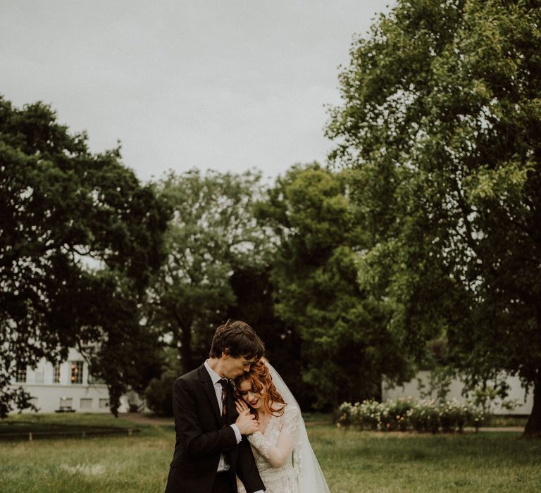 Bride wearing lace wedding dress and deep red lipstick holds the hand of her groom as they walk across manicured gardens 