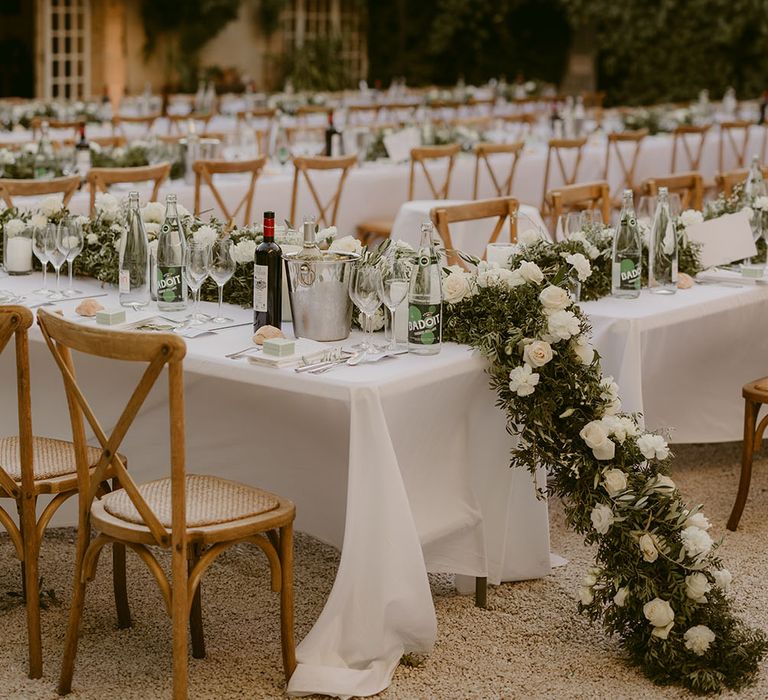 Outdoor wedding reception complete with banquet tables with white tablecloths, floral table runners and fairy light canopy hanging above