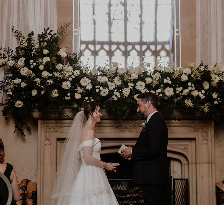 Bride and groom stand in front of a big fireplace with large white flower arrangement for classic wedding