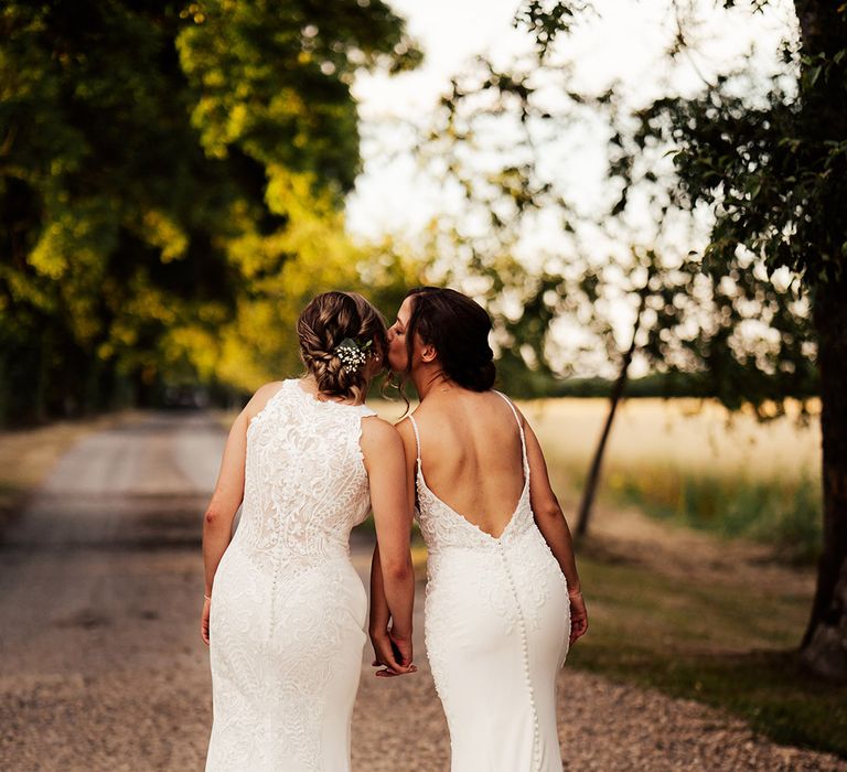 Bride leans in to kiss the bride on the cheek as they both wear lace wedding dresses with button detail on the back 