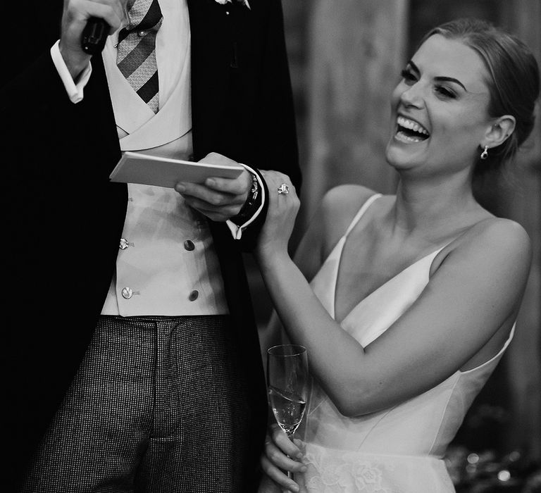 Groom reads out his speech as the bride laughs and rests her hand on the groom's arm 