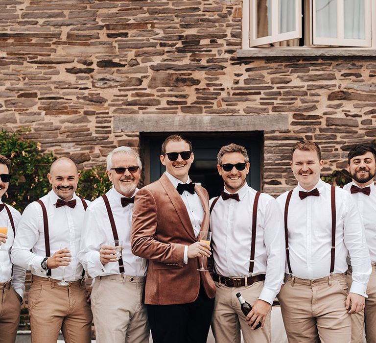 All the groomsmen in white shirts, burgundy braces and bow ties with the groom in a pink velvet suit jacket