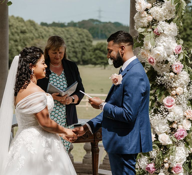 Bride & groom read wedding vows during ceremony outdoors surrounded by floral archway 