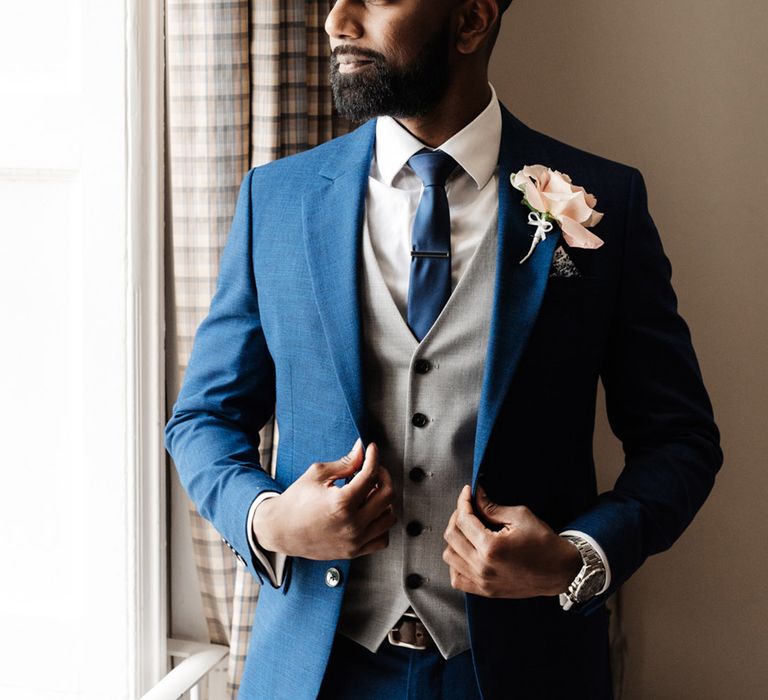 Groom wears blue suit with grey waistcoat and pastel pink floral buttonhole 