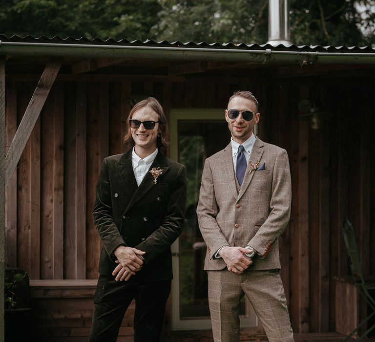 Groom wearing green suit and dried floral buttonhole stands beside wedding guest in grey suit 