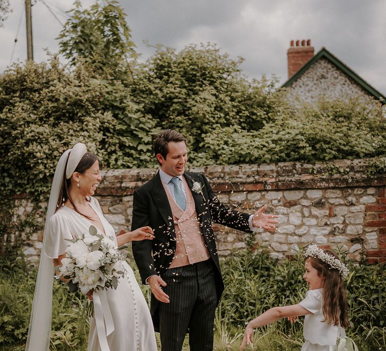 Flower girl in a white dress with white flower crown smiling with the bride and groom throwing confetti 