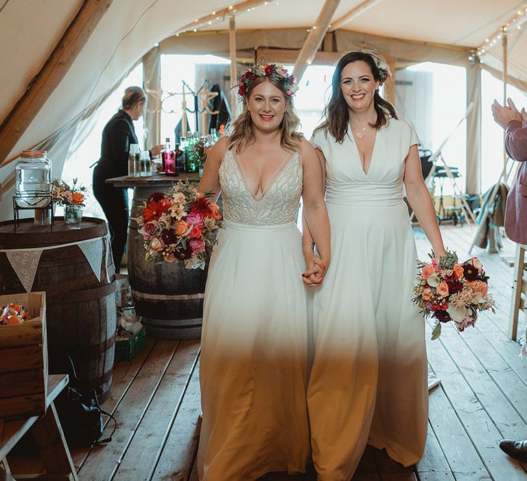 Brides walk hand in hand into the tipi for their wedding reception as guests stand and applaud them 