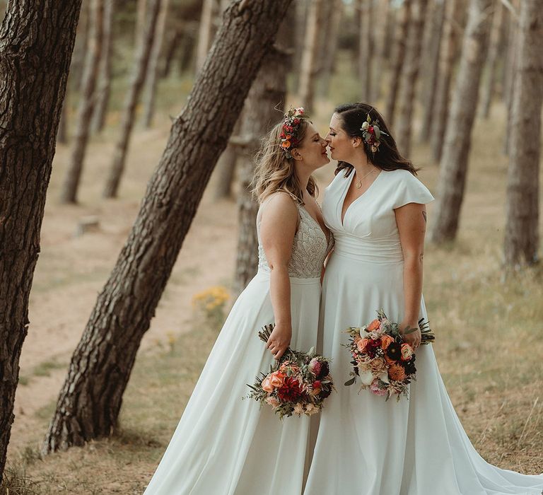 The two brides lean in for a kiss as they stand in the woodland 