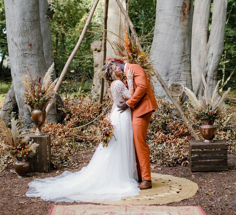 Bride nad groom hugging in the forest at harvest wedding with triangular wooden wedding arch and bronze flower pots with dried autumnal floral arrangements with white dried cotton stems, terra cotta celosia seeds, green foliage