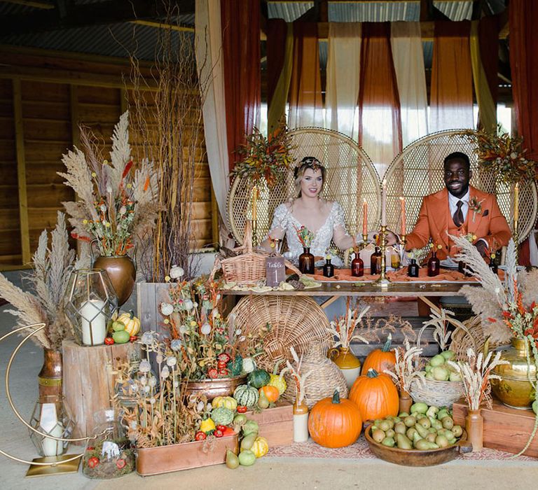 Bride and groom at autumnal harvest themed wedding tablescape with orange and yellow tapered candles, pumpkins, gourds, vegetables, dried flowers, pampas grass, gold sustainable decor and seed cards to add different textures and tones