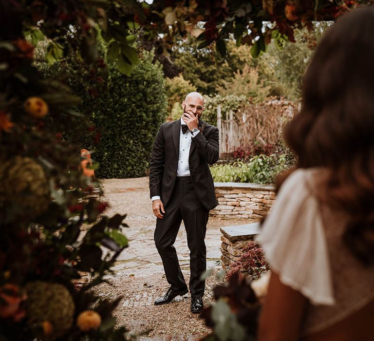 First look moment; Groom in classic black tuxedo with black bowtie looking at bride in open-back detail lace wedding dress