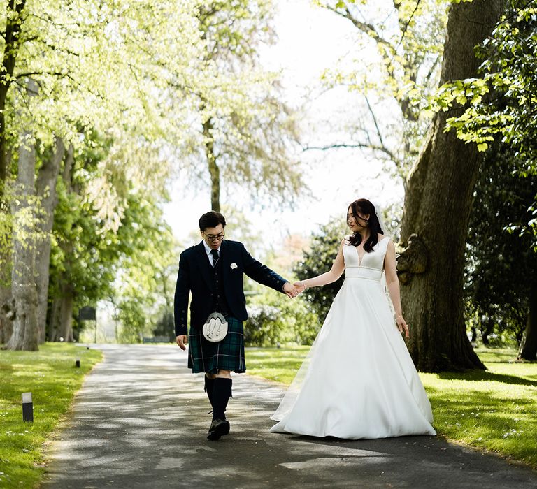 Groom in tartan green kilt walks alongside his bride in Princess dress with embellished waistband 