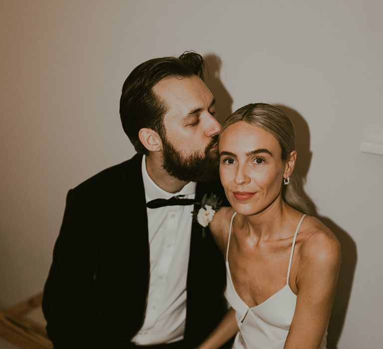 Bearded groom in a black tuxedo kisses the bride who looks into the camera with sleek ponytail hairstyle in a fitted wedding dress 