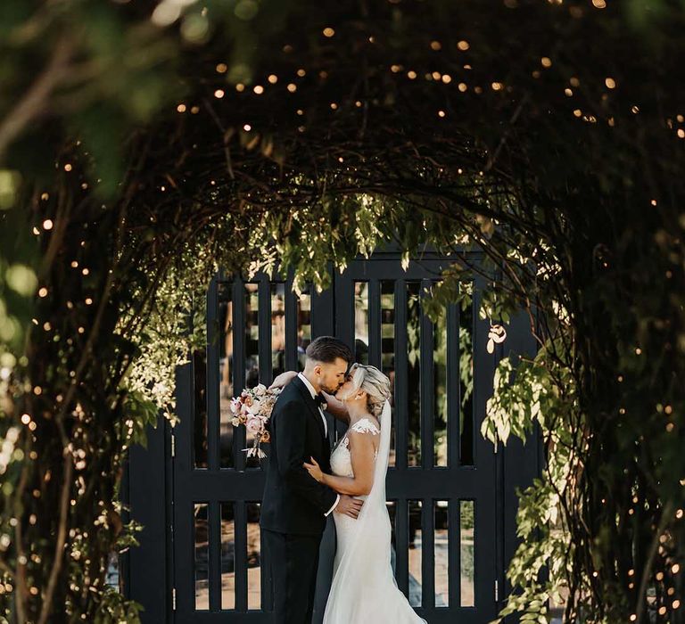 Groom in classic Ralph Lauren tuxedo and bride in lace wedding dress with sheer, lace top and clusters of lace flowers on the shoulders and plunge neckline kissing at Old Kent Barn wedding venue 