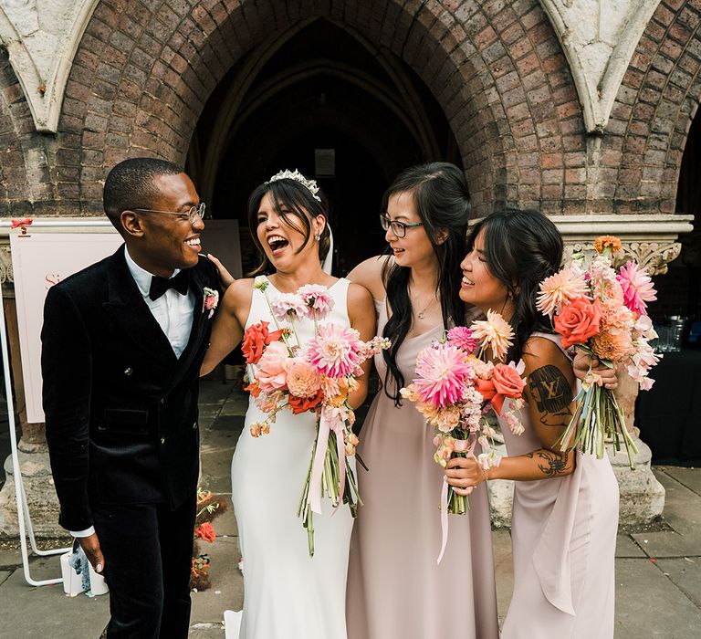 Bride stands with her bridesmaids in pale pink different styled Rewritten bridesmaid dresses and bridesman in black velvet suit 