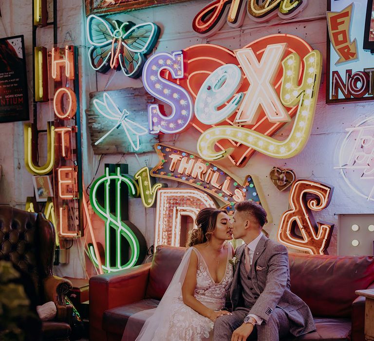 Bride in sleeveless plunge lace wedding dress  and cathedral length embroidered veil sitting on sofa with groom in grey three piece wedding suit with yellow suit and yellow pocket square below large colourful neon signs 