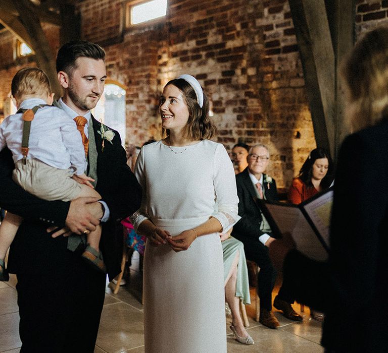 Bride in high neck wedding dress with three quarter length sleeve with the groom in a three piece suit with orange tie holding their toddler 