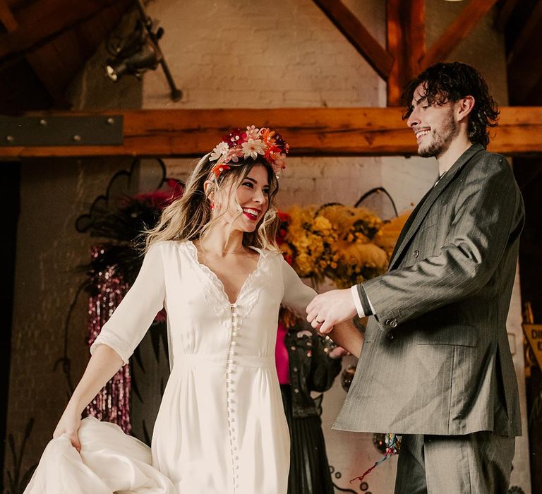 Bride in three quarter length sheer sleeve wedding dress with button and cinched waist details, pink bridal flower crown and pink fluffy open-toe strappy heels doing jumping the broom wedding tradition with groom in dark grey pinstripe suit and dr Martens boots 