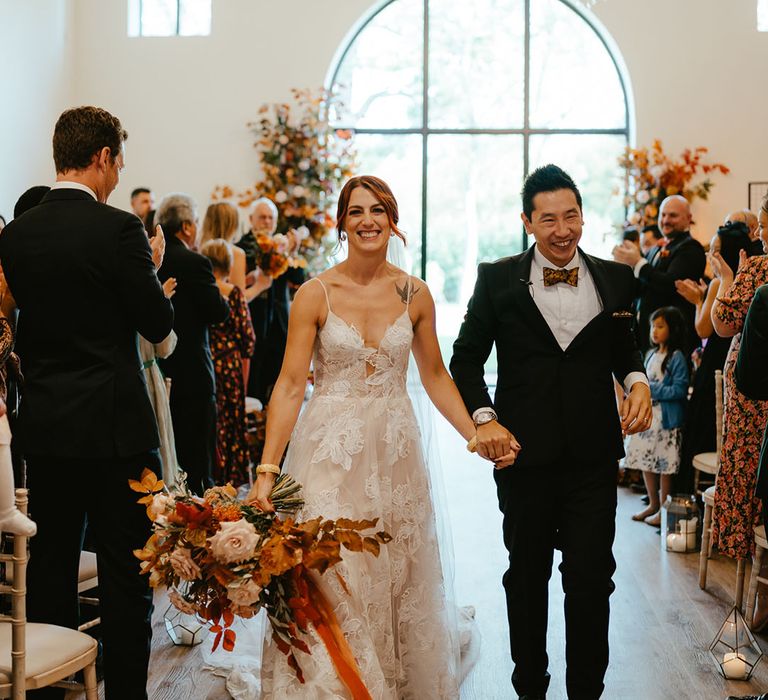 Bride holds large burnt orange bridal bouquet and wears Madi Lane Bridal wedding dress as she walks alongside her groom in black tie 