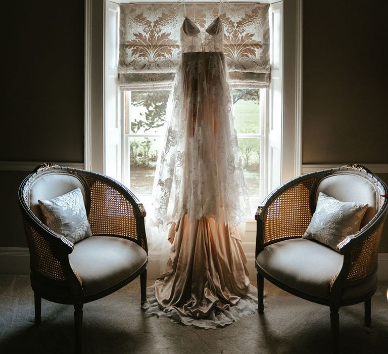 Lace Madi Lane Bridal wedding dress hangs in front of window on the morning of wedding day