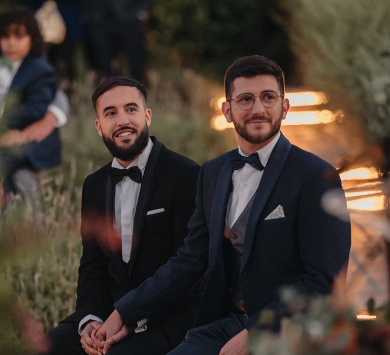 two grooms in a navy and black tuxedo holding hands during their outdoor wedding ceremony 