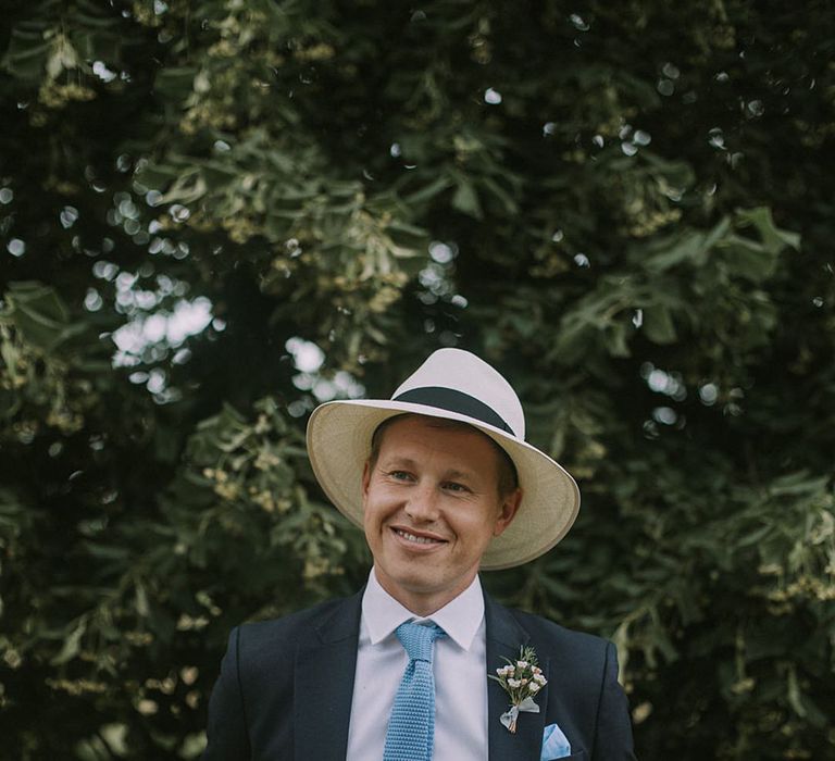 The groom stands in a dark suit with bright baby blue tie and pocket square with a white hat accessory 