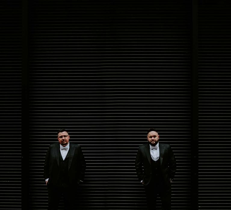 Grooms in matching three piece black tuxedos with off white bow ties and white garden rose, lavender twig and dried foliage boutonnieres doing couples wedding photoshoot at The Gherkin wedding venue London