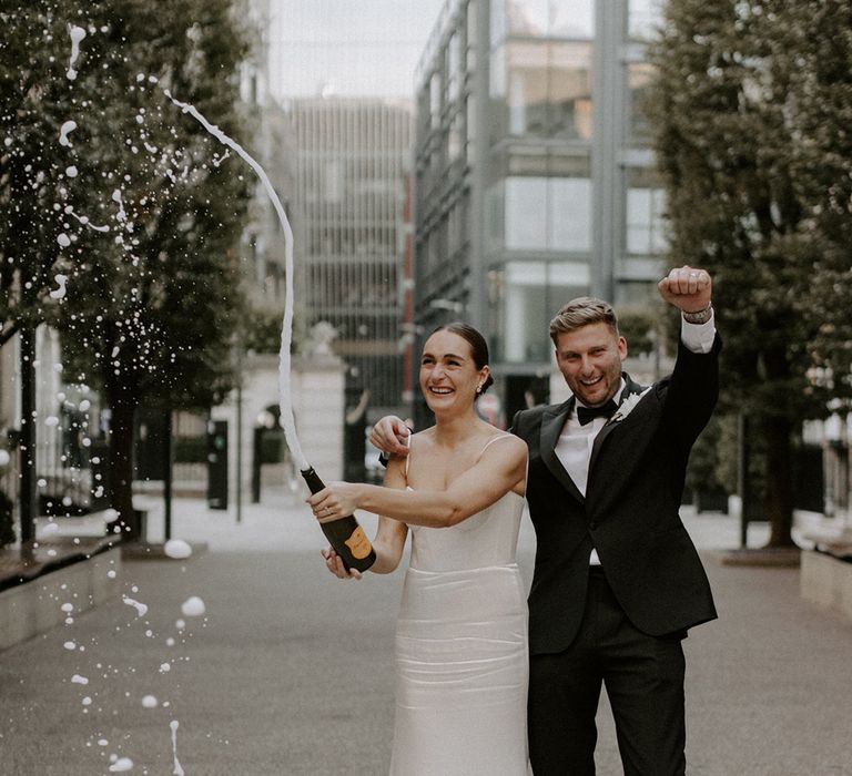 The groom in black tux pops champagne with the bride to celebrate their wedding 