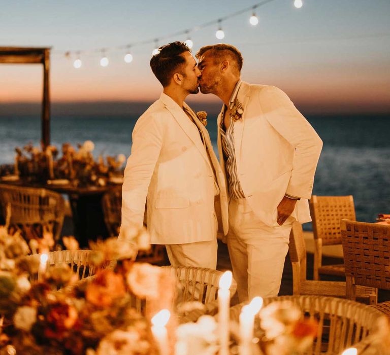 Groom in mens white wedding suit kissing groom in matching suit under suspended fairy light wedding lighting by rustic burnt orange wedding tablescapes 